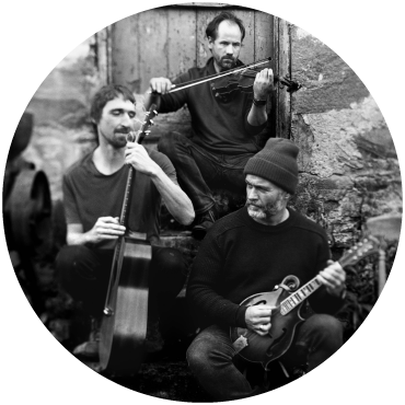 The Langan Band trio sit in front of the steps of an old building playing a fiddle, a bass guitar, and strumming a guitar. The photo is black and white.