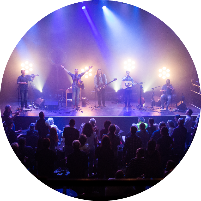 The band members stand on stage in front of a packed audience, bathed in blue and white lights