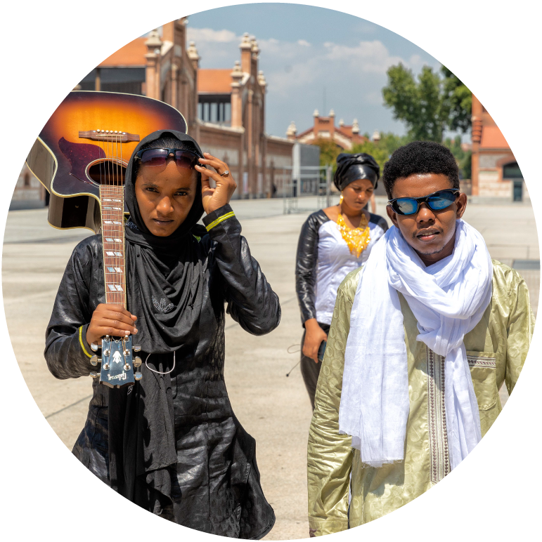 3 members of Les Filles de Illighadad are walking down a street. They wear robes and scarves. One carries a guitar on her shoulder. 
