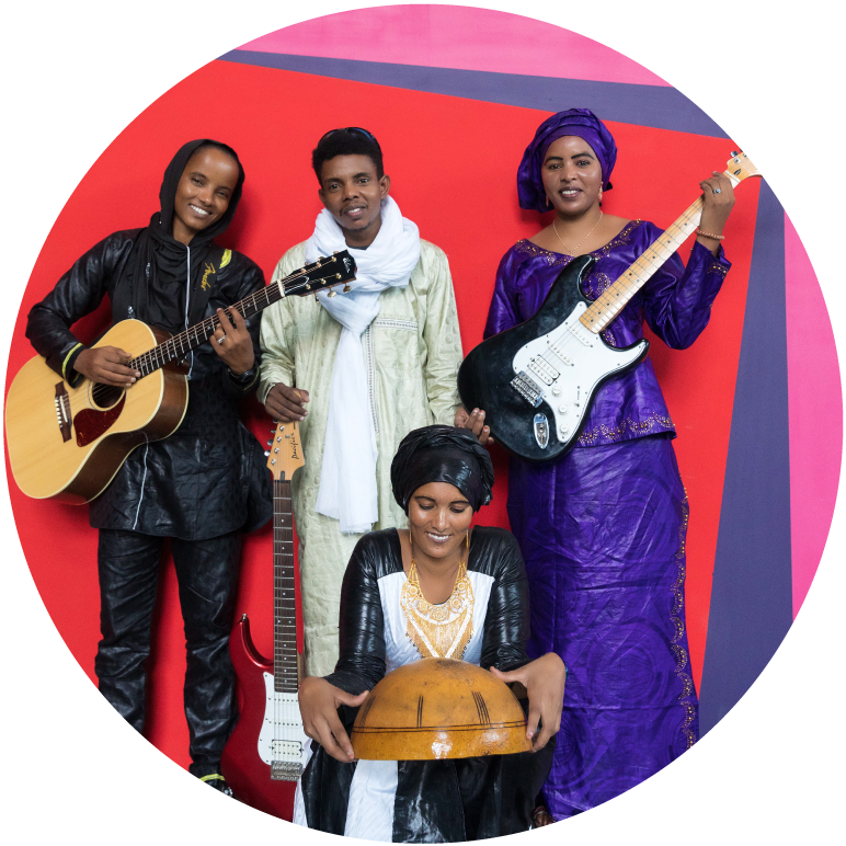 4 members of  Les Filles de Illighadad stand in front of a red and blue painted wall. 3 hold guitars and the other poses with a drum.