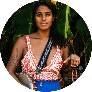 Leyla McCalla smiles into the camera with her brown hair down over her left shoulder. Their is a gray banjo strap over her left shoulder, and her right hand is resting on her banjo. She's wearing blue bottoms and a red and white patterned top.