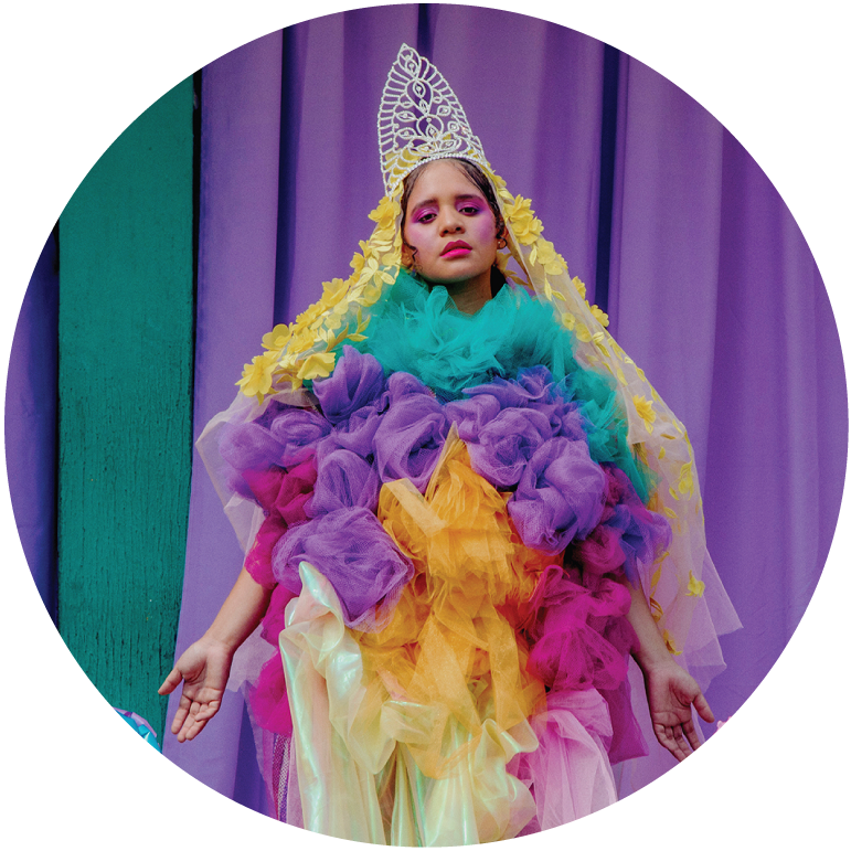Lido Pimienta wears a multicolor highly textured outfit with a long blonde wig and silver tiara