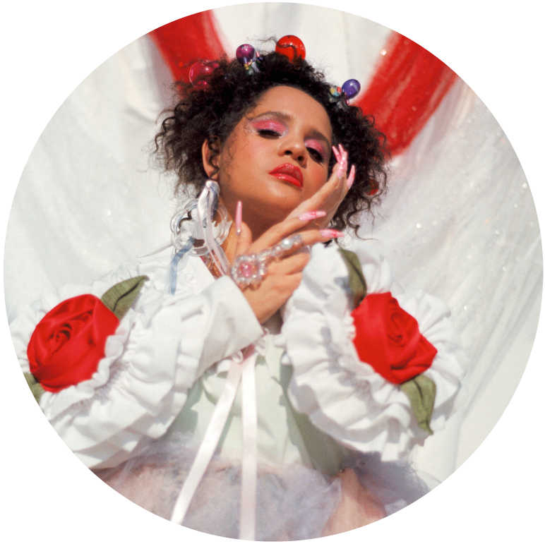 Lido Pimienta wears a white top with big red roses on the sleeves with their hands folded across their neck and their left hand rests against their cheek