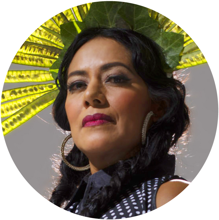 A headshot of Lila Downs. She has medium-toned skin with dark braided hair. She has on gold hoop earrings, pink lipstick, a black shirt with white polka dots, and a headdress of green leaves.