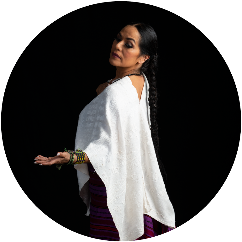 Lila Down in front of a black background. She has a medium skin tone and dark hair in a braid. She wears a white tunic, bracelets,  and looks over her shoulder 