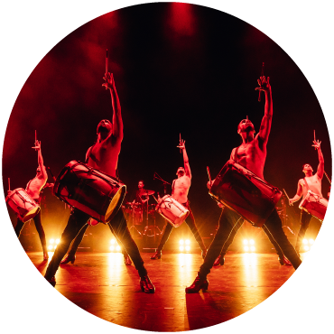 Five performers of Malevo stand on stage with their feet apart and hands extended holding mallets and drums on stage lit by red lighting.