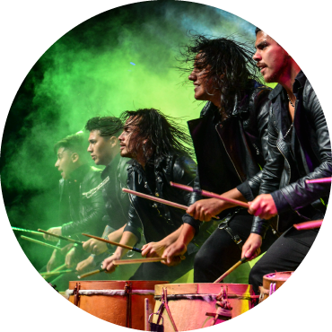 Five people with long and short black hair wear black pants and leather jackets. Green smoke is around them as they look in front of themselves and play the drums.
