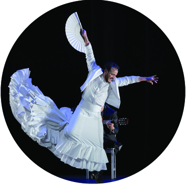 Manuel Linan dancing in a white dress with the bottom ruffles flipping upwards to the air and his arms outstretched