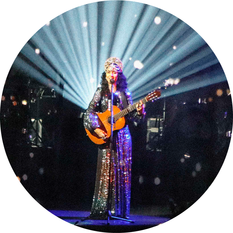 Marisa stands center stage wearing a sparkling dress and tiara. She holds a brown guitar. Spotlights shine from behind her up to the sky, creating a halo effect