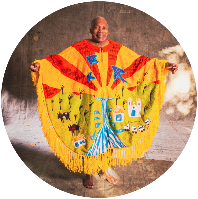 Milton stands bald and barefoot against a light brown backdrop. He wears a big yellow fabric poncho with yellow fringe along the edges and red, blue, and green detailing.