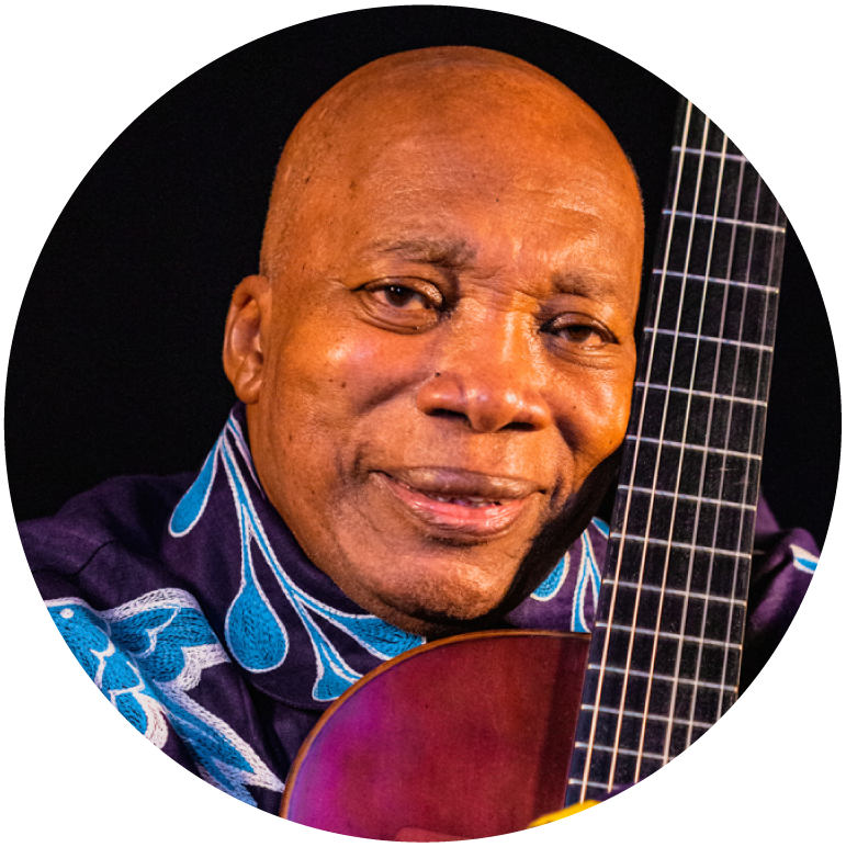 Milton has a shaved head and a smile on his face, hugging his guitar pulled close to his body with the neck of the guitar touched up against his right cheek smiling at the camera
