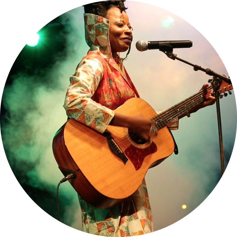 Natu Camara stands in front of a green background and white fog, while playing an acoustic guitar and singing into a microphone. She has dark skin and black hair that is in an updo. She wears a white, green, and orange jacket.