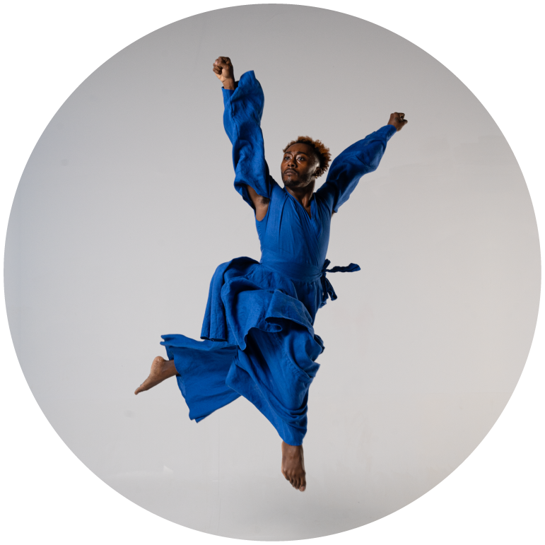 A dancer in a blue flowing jumpsuit leaps with both arms stretched above their head