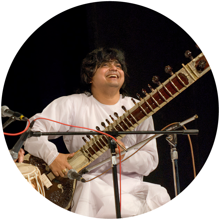 Niladri Kumar wears a white outfit holding his sitar in front of a microphone. He plays with a smile on his face.