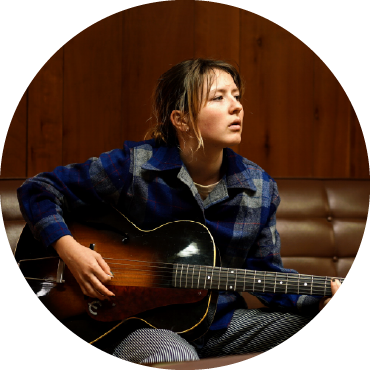 Nora Brown gazes off into the distance and strums a guitar as she sits on a brown sofa