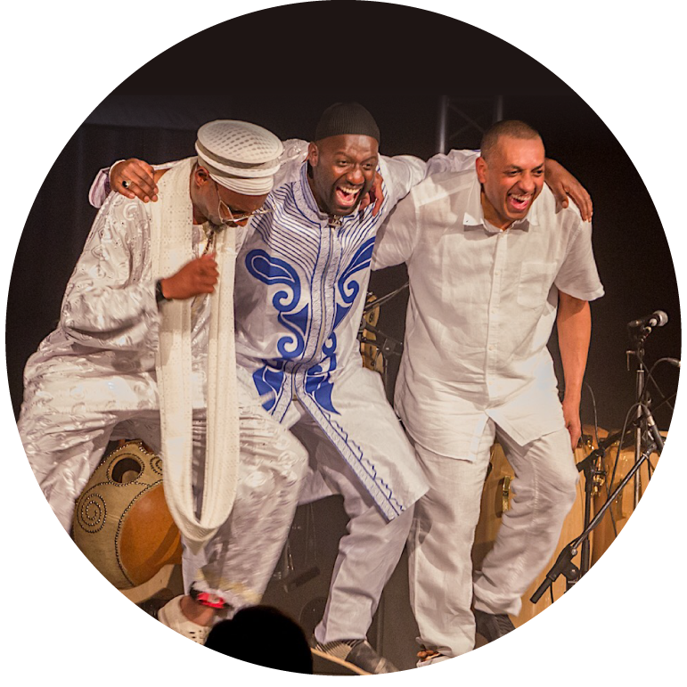 Three musicians dance on stage, arms around each other in a line, dancing wearing all white