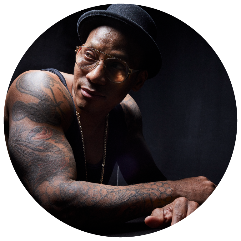 Pedrito Martinez leans forward with his arms crossed wearing a black tank top, black hat, and sunglasses