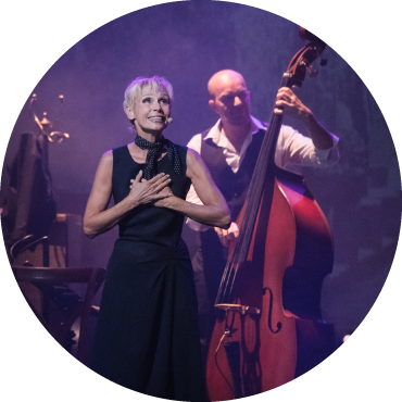 Nathalie Lermitte wears a black gown and is captured singing live on stage with a cellist playing in the background
