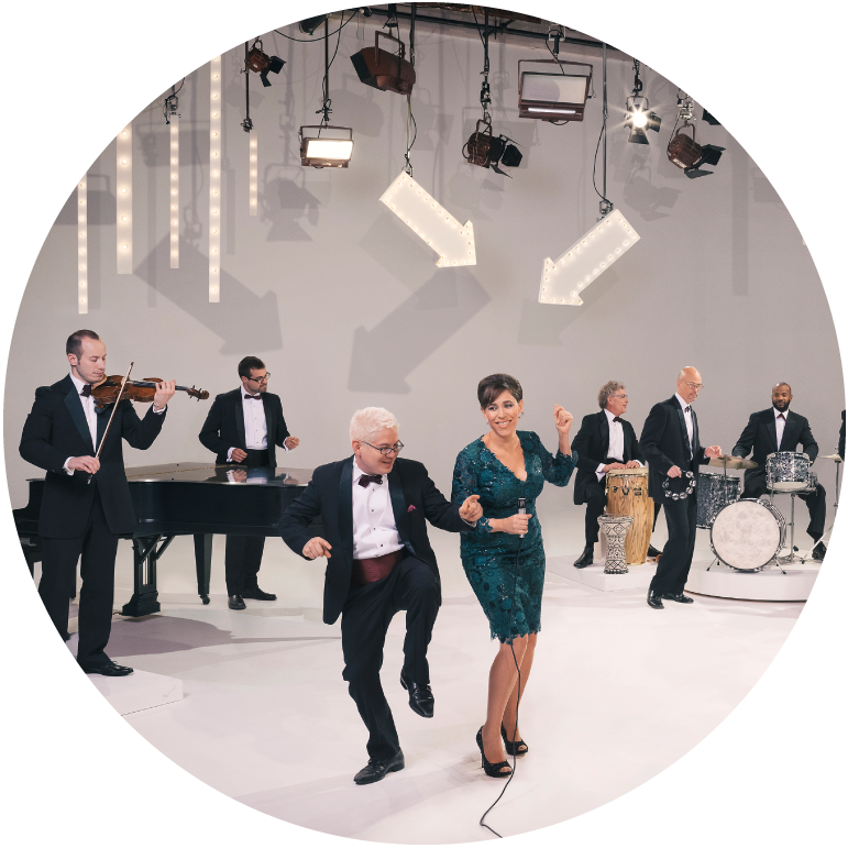 China Forbes is in a green knee-length dress and Thomas Lauderdale is in a black suite with a white shirt. The band plays around them against a gray backdrop with two large white arrows hanging from the ceiling