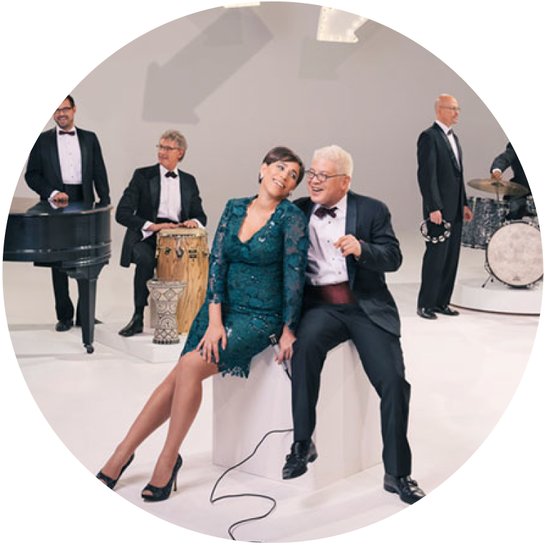 China wears a green evening dress and Thomas wears a black suit. They sit in the center on a white block against a gray background with members of the band in the background