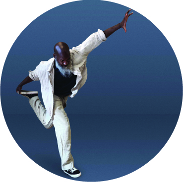Raphael Xavier wears a white suit with a black undershirt against a blue background. He stands on one foot with his left foot held behind him with his right hand and his left hand extended into the air