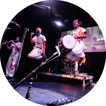 Red Baraat band members are on stage holding various instruments and are all wearing white jumpsuits with flowers painted on. The two members in the center are jumping.