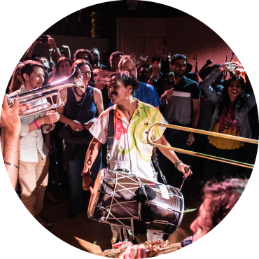 Red Baraat band members are standing in front of an audience, all wearing white jumpsuits with designs painted on. The person in the center of the circle of people is smiling and wearing a drum.