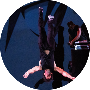 A person wearing all black is upside-down mid-air doing a flip. People sitting on the floor to the left are wearing all black, while another group of people on the opposite side are wearing all red.