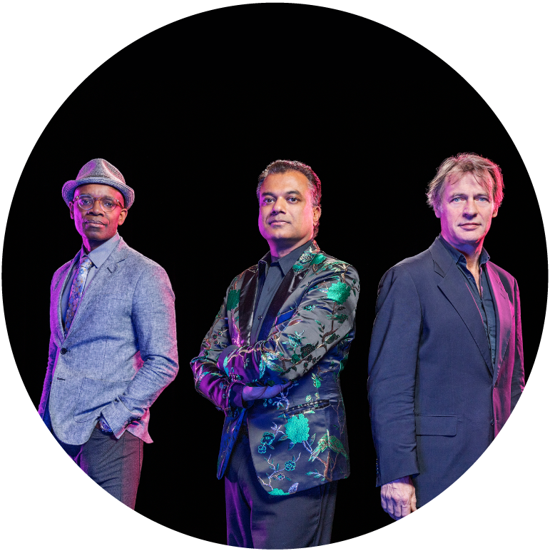 Rudresh stands in the middle between two men. On his right, a dark-skinned man with a gray suit and hat who has his hands in his pockets. The man on the left has light skin and hair and wears a blue suit. Rudresh has his arms crossed and wears black shirt and pants and a flowered jacket. All three look at camera.