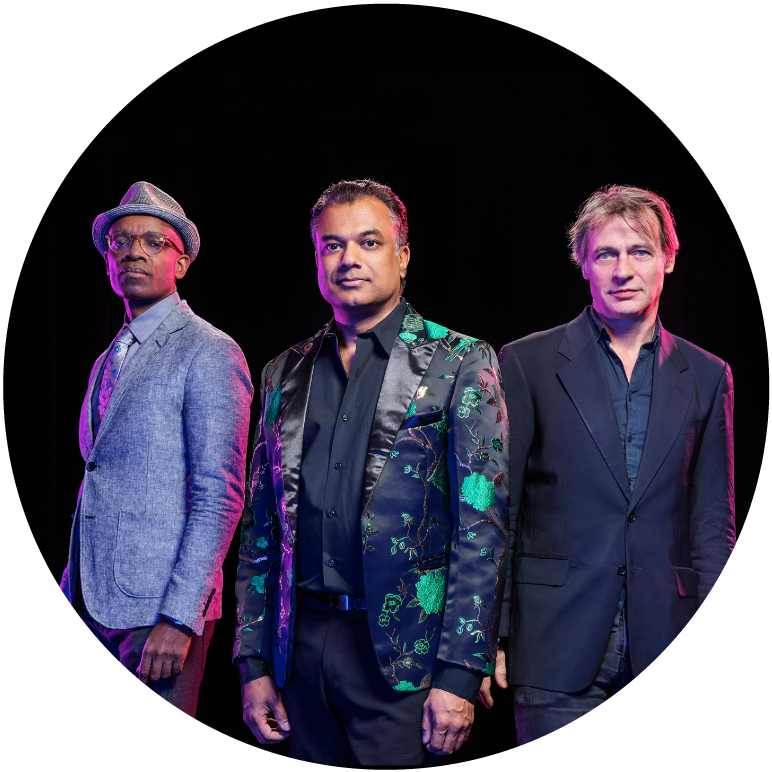 Rudresh stands in the middle between two men. On his right, a dark-skinned man with a gray suit and hat who has his hands in his pockets. The man on the left has light skin and hair and wears a blue suit. Rudresh wears black shirt and pants and a flowered jacket. All three look at camera.