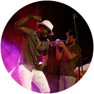 Sidy Samb dances on stage in a cheetah print shirt and white fedora. A performer plays the trumpet in the background.