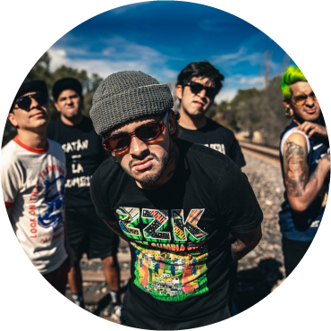 Son Rompe Pera members wear graphic t-shirts and pose together by train tracks