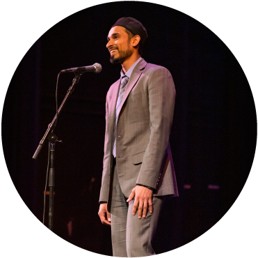 A circular image of a person in a gray suit. They are standing in front of a microphone and smiling. 