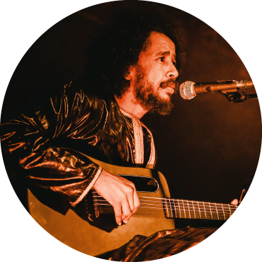 A member of Tamikrest is sitting and singing while playing the guitar. They have medium length curly black hair and are wearing a patterned shirt.