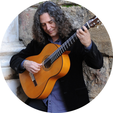 Tomatito wears a black blazer and plays guitar in front of a brick wall
