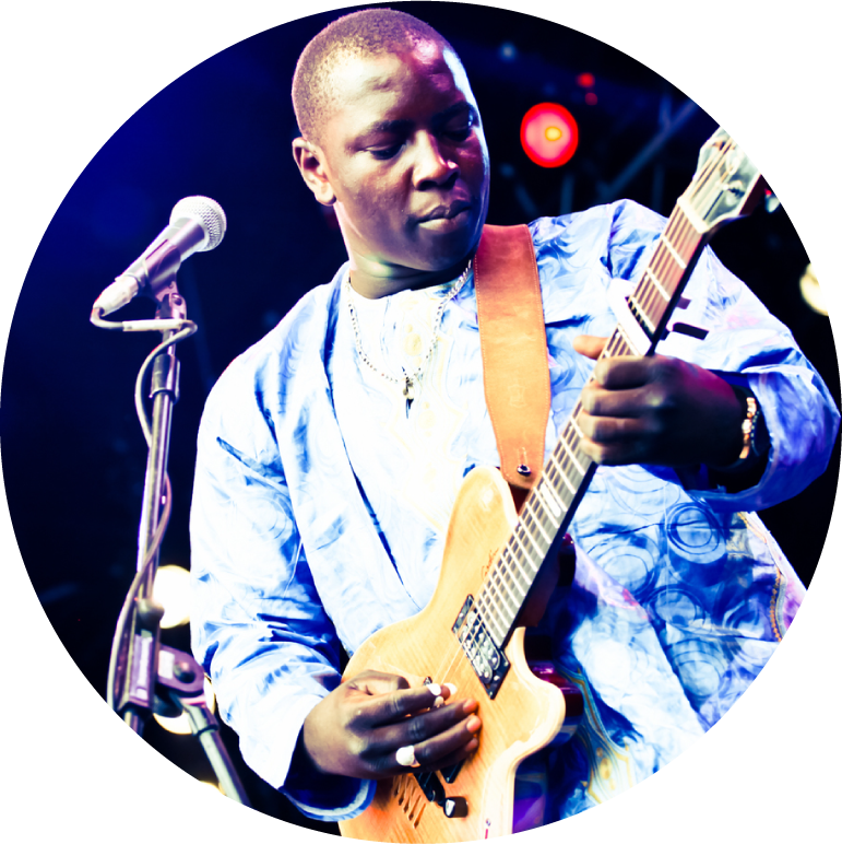 Vieux Farka Toure wears a blue and white shirt. There is a microphone in front of him, as if he is performing on stage. He holds his guitar and is looking down at the guitar.