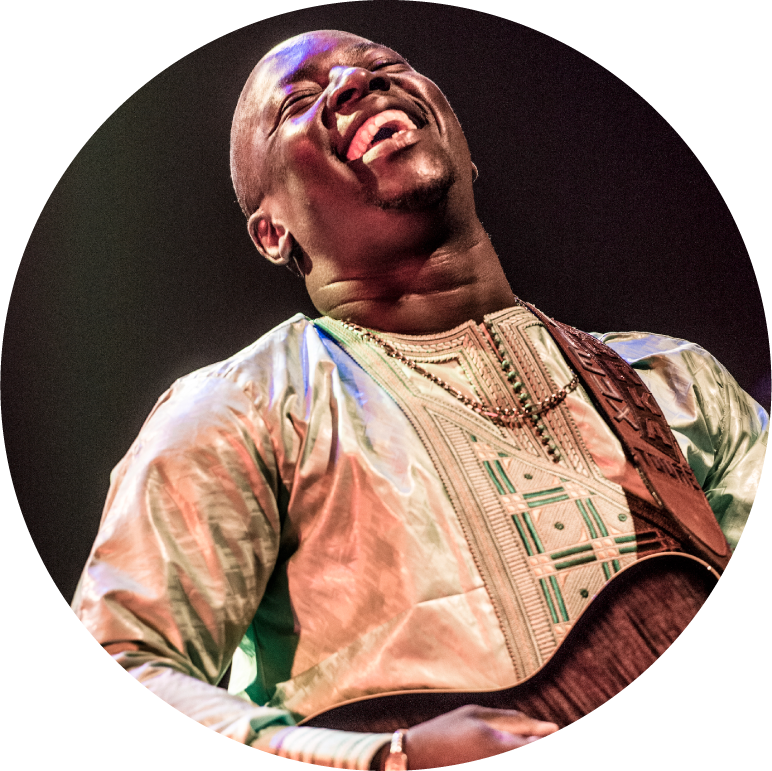 Vieux Farka Toure performs on stage against a black background. His guitar strap sits on his right shoulder and his left hand strums the guitar. His eyes are closed and he is smiling. 