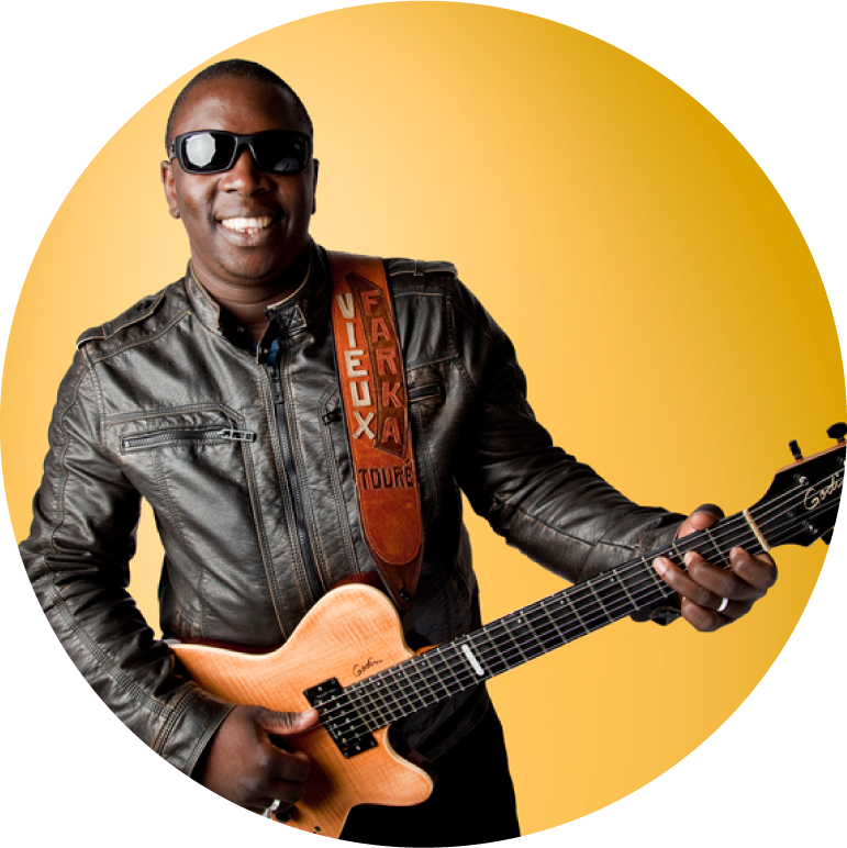 Vieux Farka Toure wears black sunglasses and a black leather jacket. He is holding his guitar and smiling against a solid yellow backdrop.