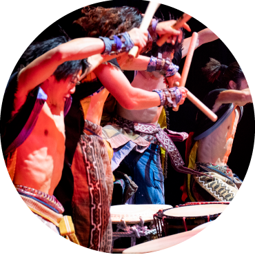 A circular image of a group of four men wearing open black shirts and varying colors of pants are hitting drums in front of each of them.