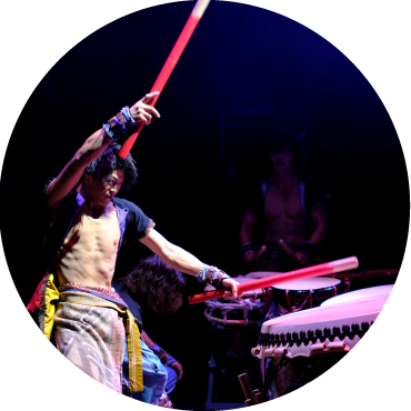 A circular image of a man wearing an open black shirt and multi-color pants is holding large red drum sticks. His right arm is raised, ready to strike down on the large drum in front of him.