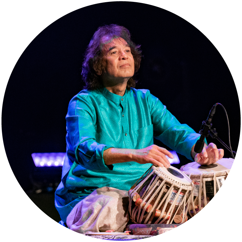 Zakir Hussain wears a teal top and he plays his tabla sitting down, eyes closed
