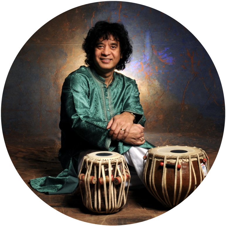 Zakir sits on the floor in front of a gray background and behind tabla drums. He wears a teal tunic.