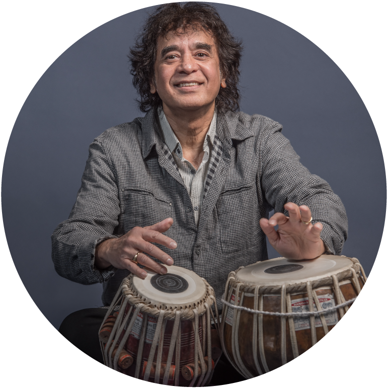 Zakir sits behind tabla drums in front of a gray background wearing a gray shirt