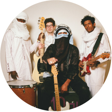Mdou Moctar band holding their instruments and sitting against a white wall. 