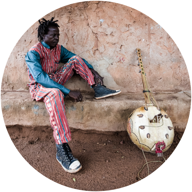 Baba Commandant wears blue and red clothing and is pictured sitting outdoors with an instrument