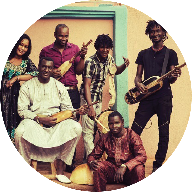 6 members of Ngoni Ba stand and sit with their instruments against a white building with a mint green door frame