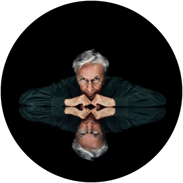 Caetano Veloso looks towards the camera with his arms under his chin, against a black backdrop