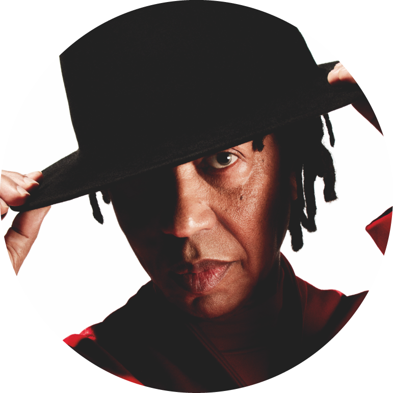 Djavan wears a black wide rimmed hat and a red shirt. He looks at the camera and has both hands on the side rims of his hat, pulling it down over his right eye