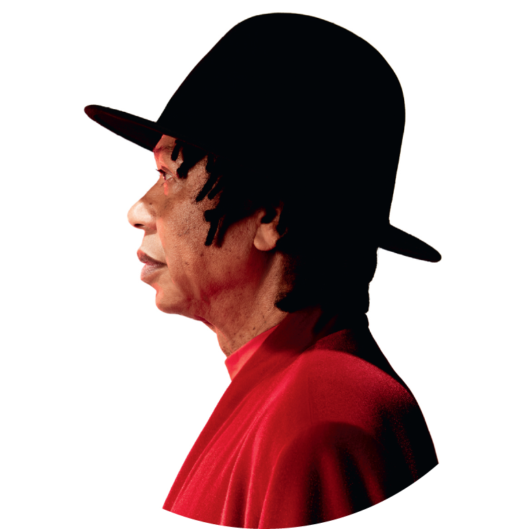 Djavan wears a black hat with a large rim and wears a red shirt. He stands sideways, staring straight ahead into the distance.
