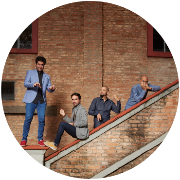 Harold López-Nussa's quartet pretends to play instruments by the stairs of a brick building. Two men are on top of the staircase posing, two men are behind the staircase wearing blue shirts.